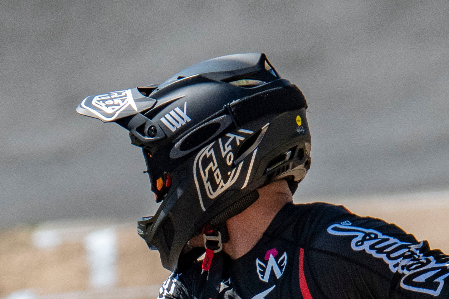 A BMX rider in full gear and helmet, marked with number 10, prepares to start a race on a ramp under a metal roof.