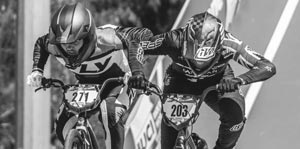 Two cyclists wearing helmets and racing gear compete closely in a BMX race, both numbered bibs visible: #271 on the left and #203 on the right. Black and white image.