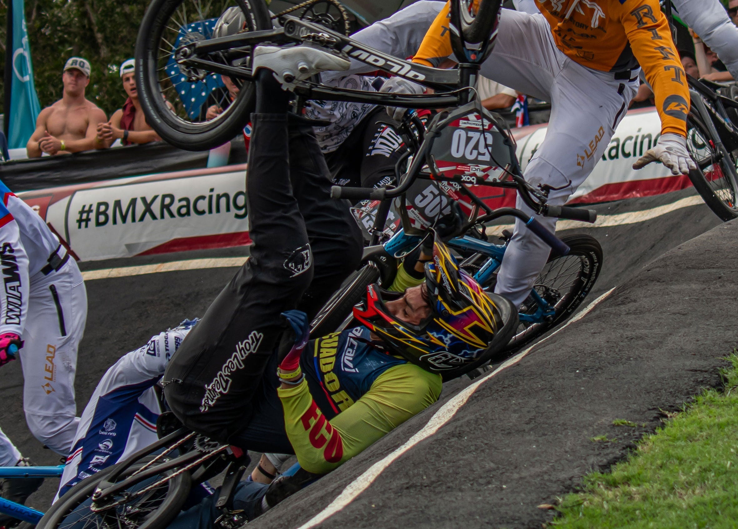A BMX rider falls headfirst over the handlebars during a race at a BMX track, with spectators watching in the background.