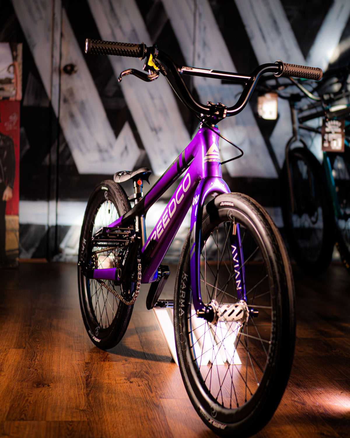 A purple SEEDCO BMX bike with black handlebars and tires is displayed indoors on a wooden floor with geometric wall art in the background.