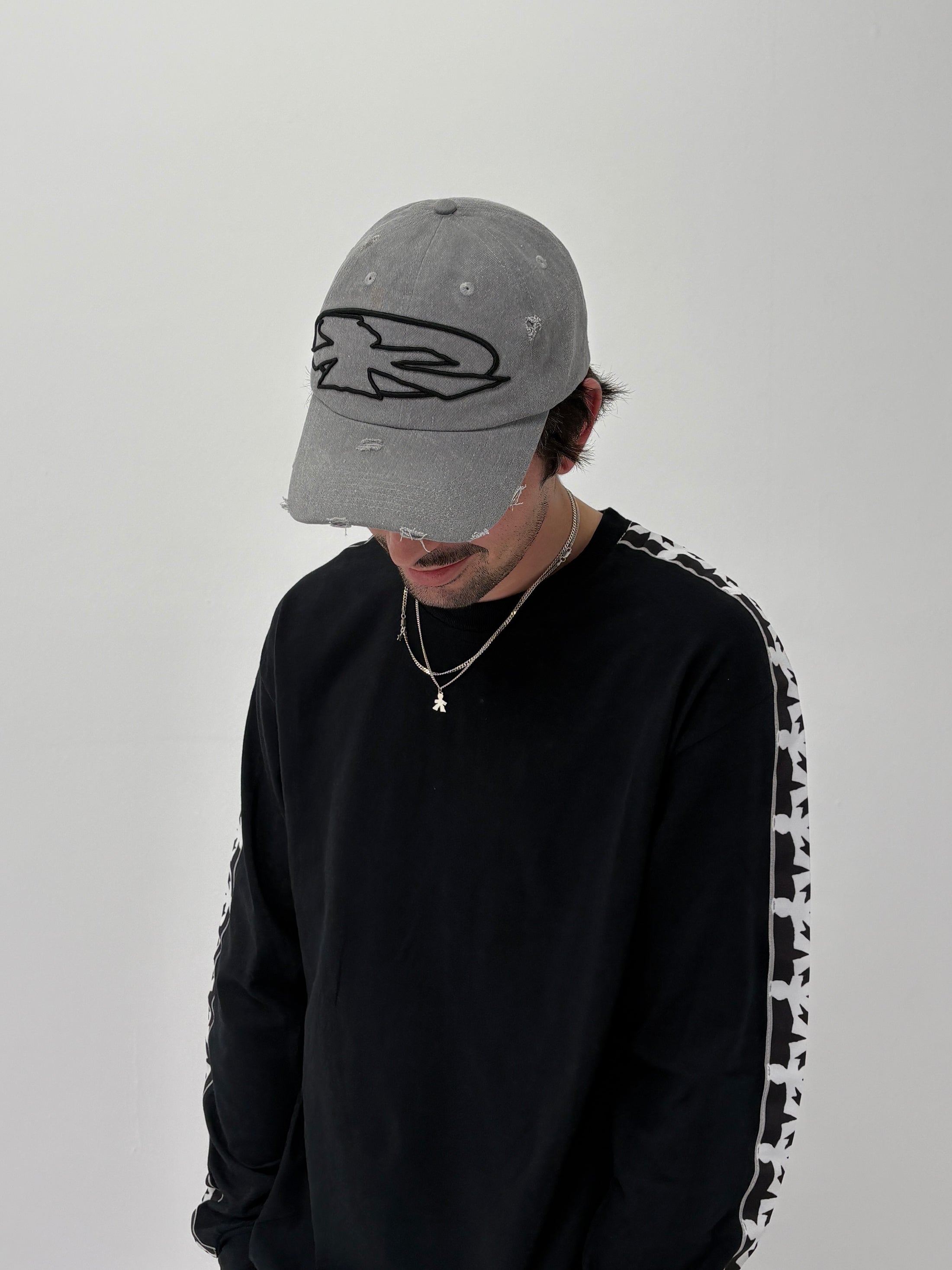 Wearing the V1da Shoreline Cap—a 100% cotton strapback with embroidered logo—alongside a black long-sleeve shirt with patterned sleeves and a necklace, this person gazes downward against a plain background.