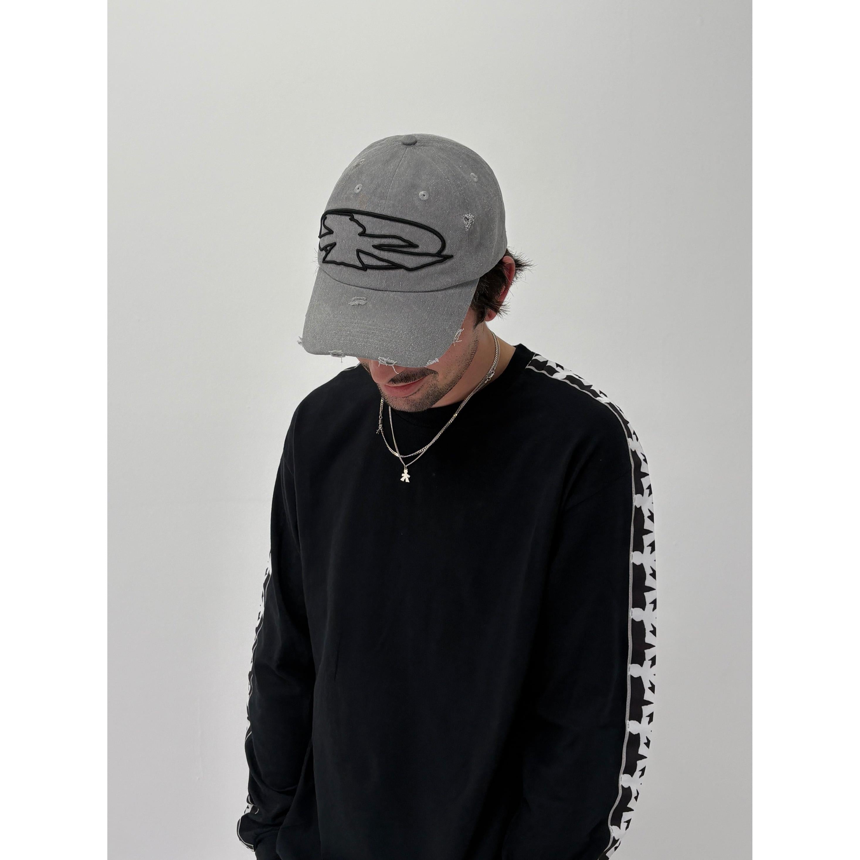 Wearing the V1da Shoreline Cap—a 100% cotton strapback with embroidered logo—alongside a black long-sleeve shirt with patterned sleeves and a necklace, this person gazes downward against a plain background.