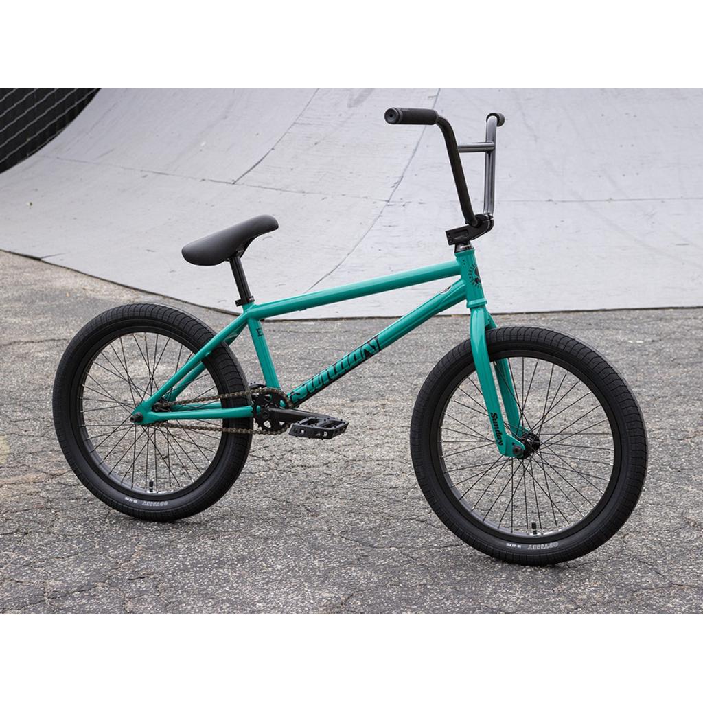 A teal Sunday EX Erik Elstran 20 Inch Bike with black handlebars, seat, and tires—featuring a full chromoly frame and Odyssey/Sunday parts—is parked on asphalt near a concrete skate ramp.