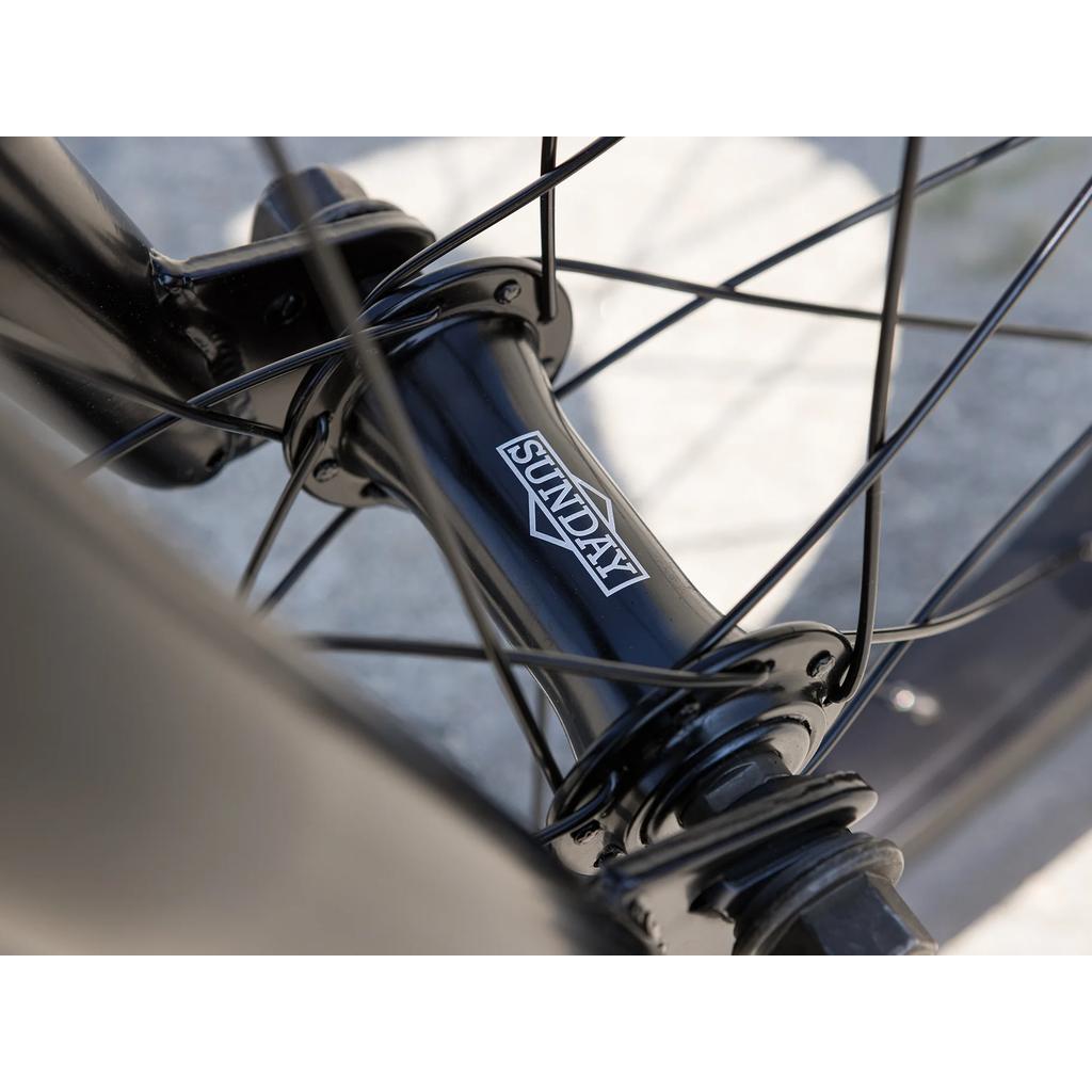 Close-up of a black wheel hub labeled "Sunday," with spokes radiating outward on the Sunday Primer 16 Inch Bike (2025). The blurred background emphasizes its pro-level BMX quality, perfect for 16” bikes.