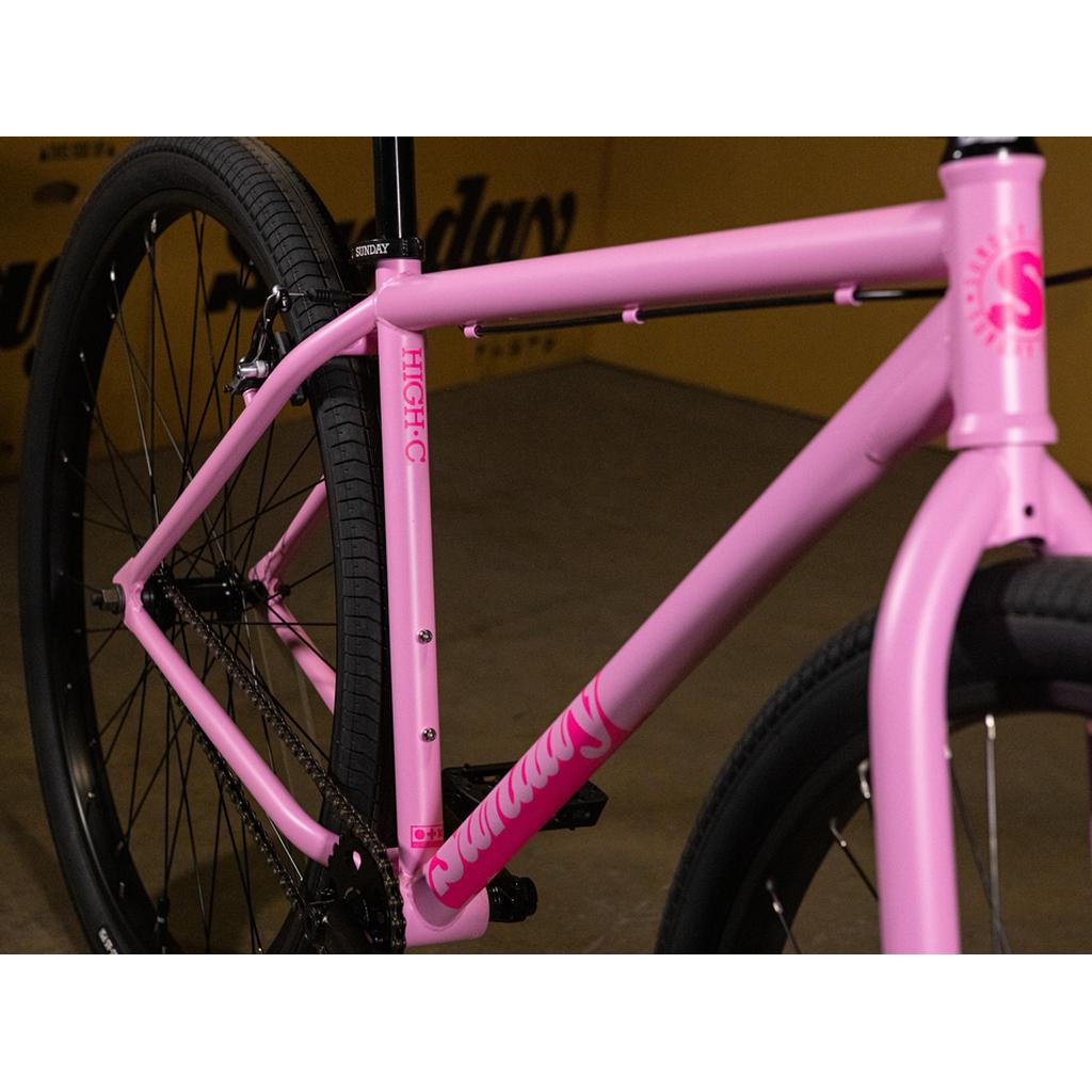 Here is a revised version using your product data: Close-up of the Sunday High C 29 Inch Bike in matte pink, highlighting its sturdy chromoly frame, rear wheel, and part of the handlebars. The bike features black tires and visible "Sunday" branding, showcasing the signature quality of this 29" cruiser.