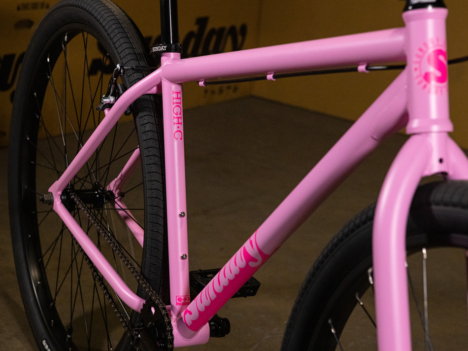 Here is a revised version using your product data: Close-up of the Sunday High C 29 Inch Bike in matte pink, highlighting its sturdy chromoly frame, rear wheel, and part of the handlebars. The bike features black tires and visible "Sunday" branding, showcasing the signature quality of this 29" cruiser.