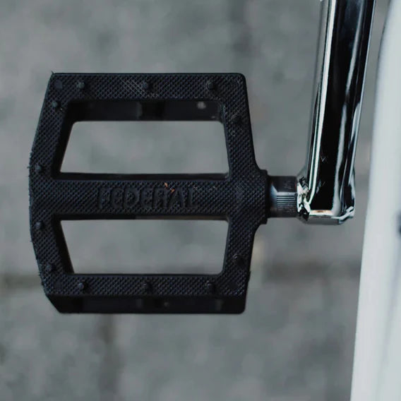 Close-up of a black Federal Contact Pedal attached to a silver crank arm, built for superior traction and grip, with a blurred grey pavement background. Federal Contact Pedals offer enhanced control for riders seeking reliability and performance.