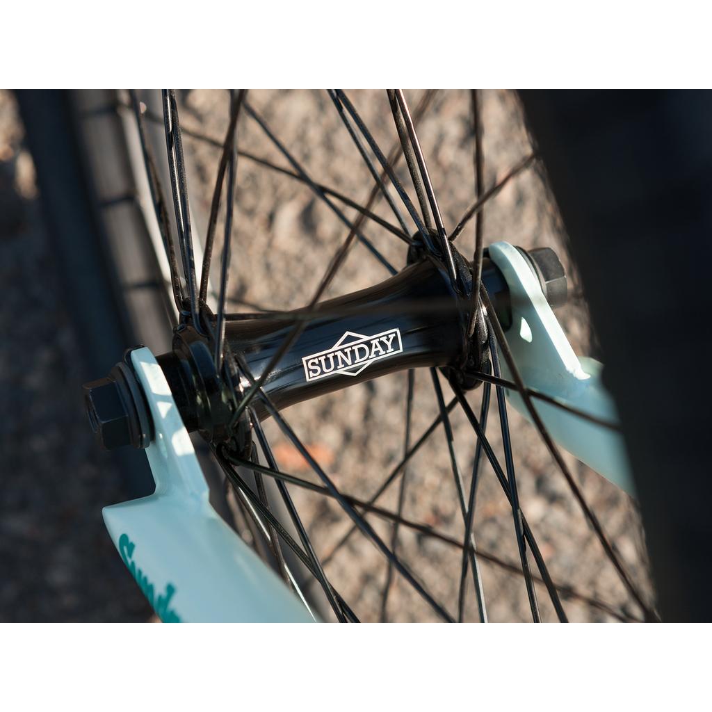 Close-up of the Sunday EX Julian Arteaga 20 Inch Bike's front wheel hub with black spokes and "SUNDAY" logo, attached to a white fork on textured pavement. Features details from Julian Arteaga’s signature bike and its 100% chromoly frame.