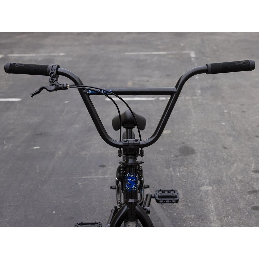 Front view of the Sunday Forecaster Maca Grasset 20 Inch Bike in black, highlighting the handlebars, brake lever, gyro cables, seat, and frame on a paved surface.