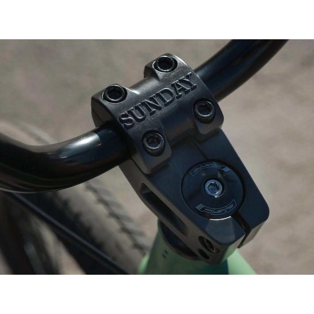 Close-up of a Sunday Scout 20 Inch Bike handlebar stem with "SUNDAY" engraved on top, sturdy bolts, and part of the front wheel—ideal for pairing with Odyssey parts for extra durability.