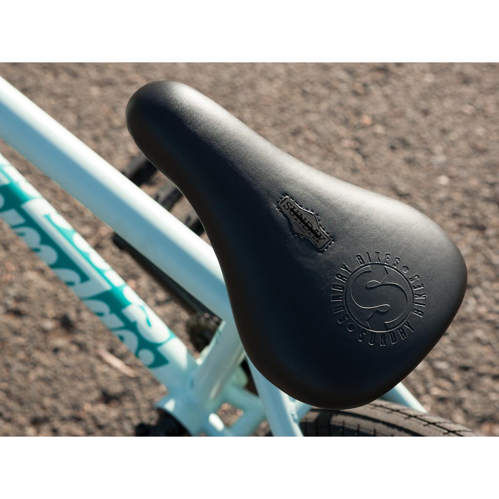 Close-up of a black bicycle seat with embossed text and logo, mounted on the 100% chromoly frame of a Sunday EX Julian Arteaga 20 Inch Bike, shown outdoors on a paved surface.