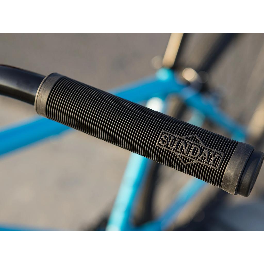 Close-up of a black ribbed handlebar grip with "SUNDAY" embossed, attached to the blue chromoly frame of the Sunday High C 29 Inch Bike (2025).