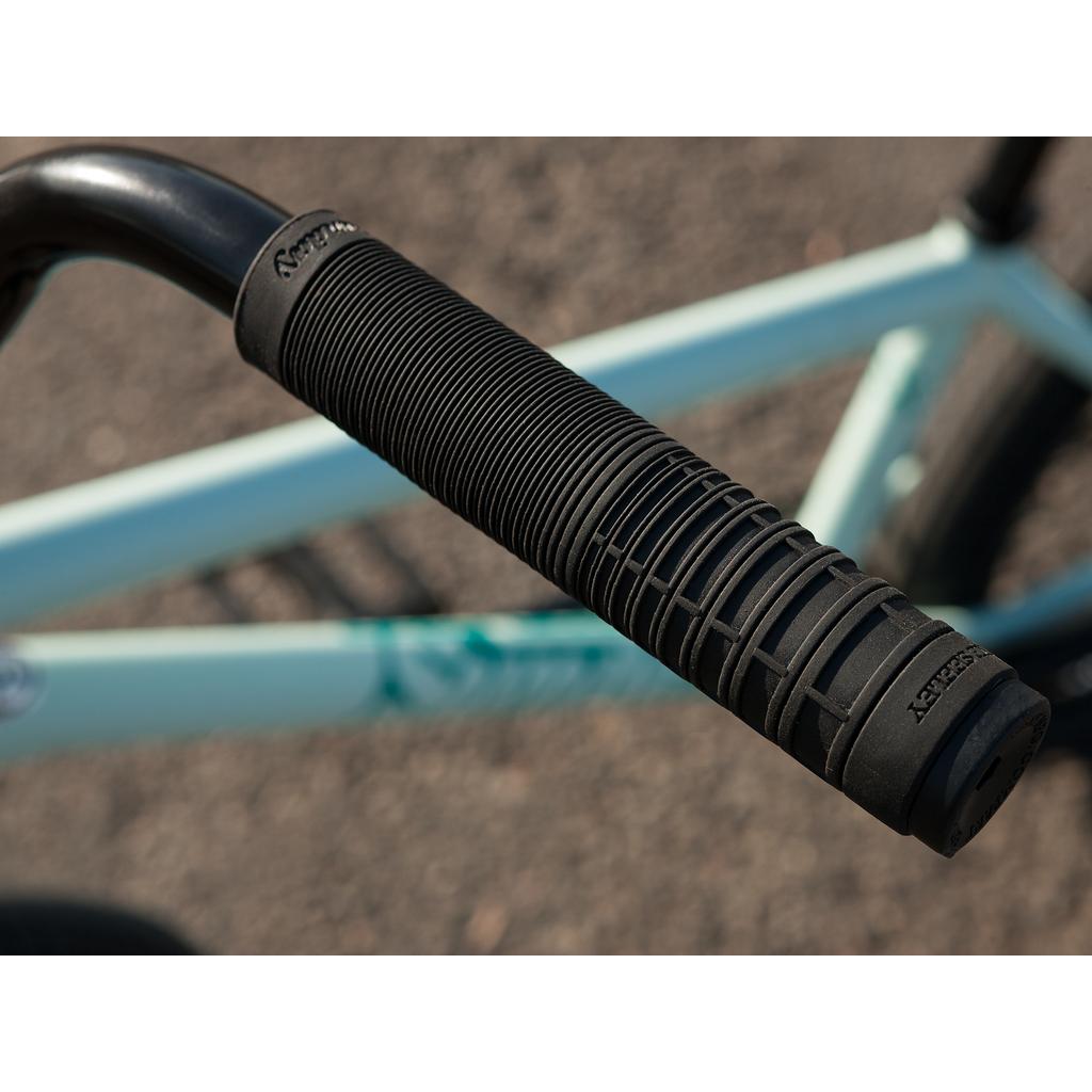 Close-up of a black rubber handlebar grip with textured lines on the Sunday EX Julian Arteaga 20 Inch Bike’s light blue frame, photographed outdoors.