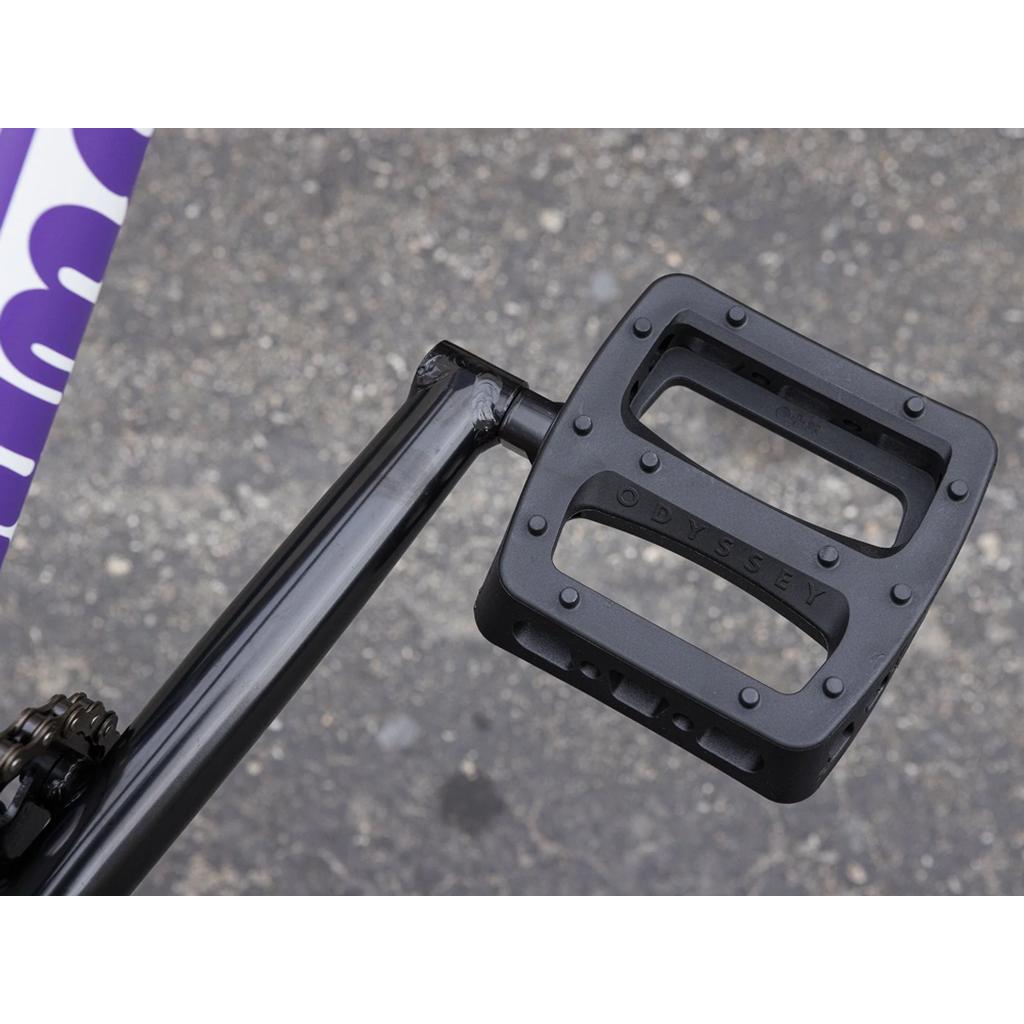 Close-up of a black Odyssey bicycle pedal mounted to the crank arm of a chromoly Sunday Forecaster 20 Inch Bike, with part of the frame and asphalt ground in view.
