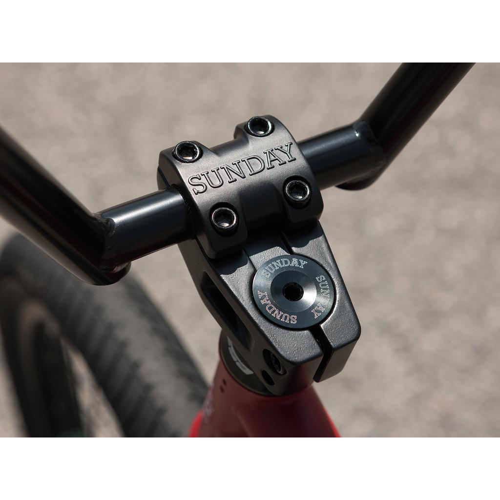 Close-up of the black BMX stem and handlebars with "Sunday" engraving, mounted on a red Sunday Forecaster 20 Inch Bike—crafted from chromoly and equipped with quality Odyssey components.