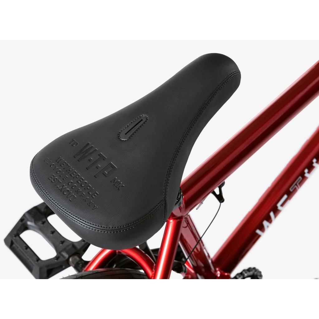 Close-up of a black bicycle saddle with embossed text, mounted on a red Wethepeople Arcade 20 Inch Bike with Salt Valon Rims, a visible pedal, and part of the bike's logo.