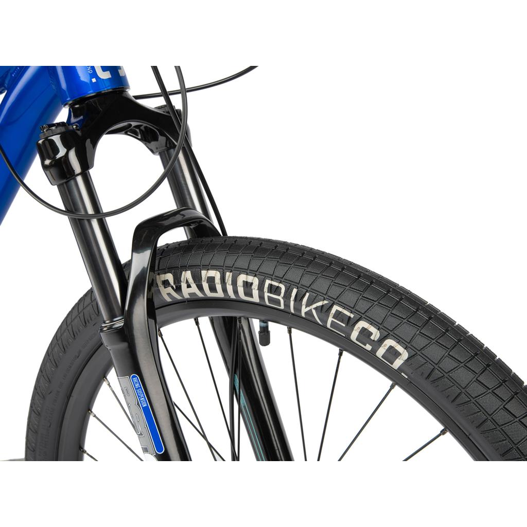 Close-up of the Radio Fiend 26 Inch Bike's front wheel with a "Radiobikeco" tire, black fork, and visible brake cables—showcasing durable 6061-T6 alloy construction for quality dirt jump performance against a white background.
