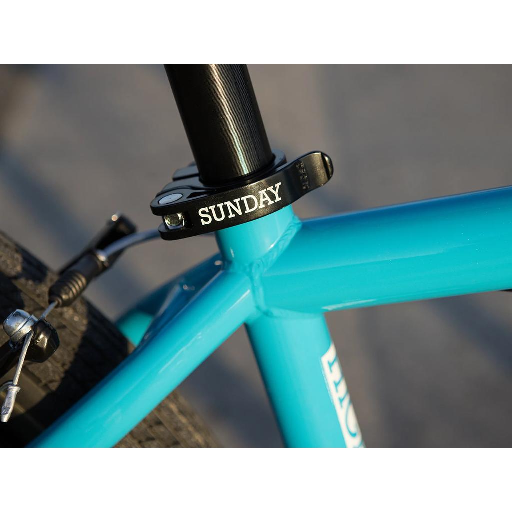 Close-up of the Sunday High C 29 Inch Bike (2025) showing its blue chromoly frame, "SUNDAY"-labeled seat post clamp, and part of the rear tire with brake cable.