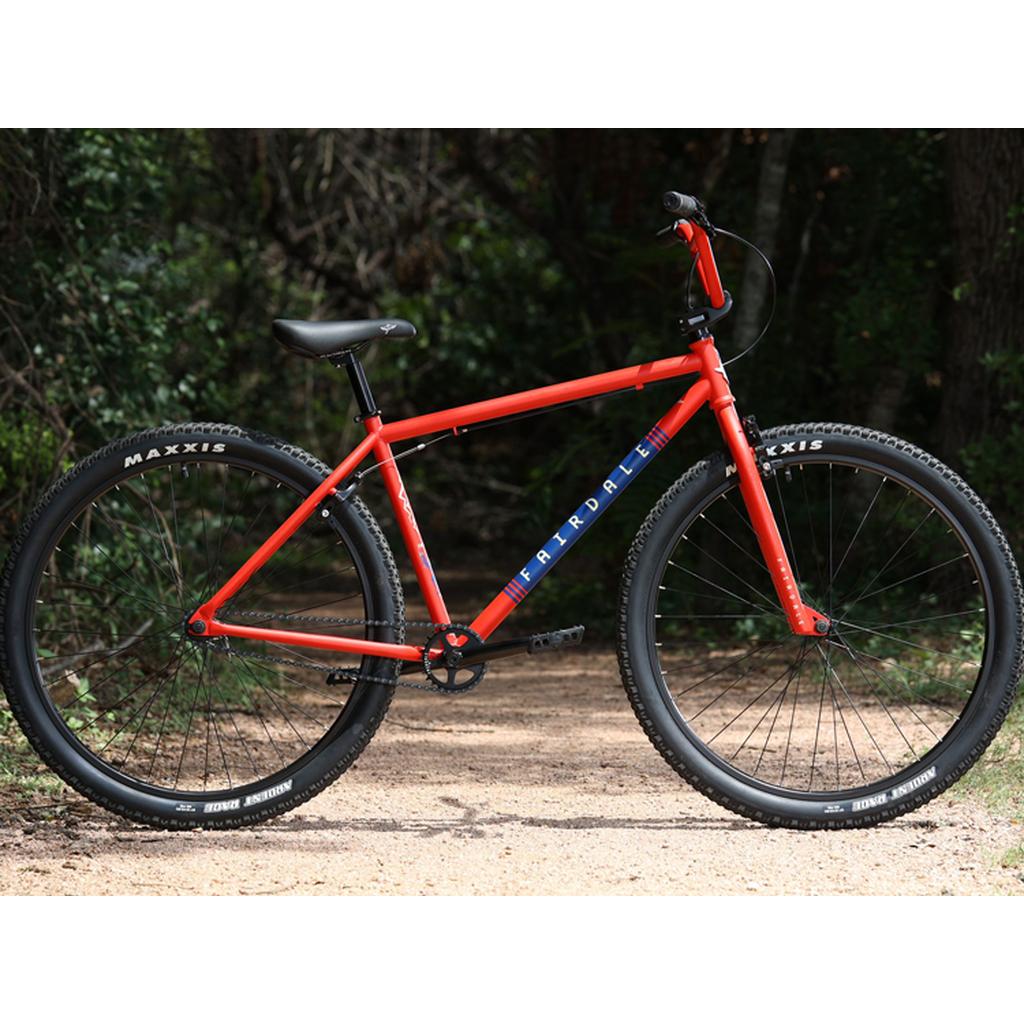A Fairdale Taj 27.5 Inch Bike (2023) with black tires and seat is parked on a dirt path among green trees, showcasing its sturdy single speed design and bold red color.