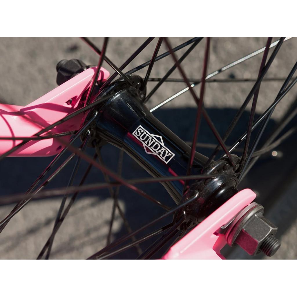 Close-up of a Sunday Scout 20 Inch Bike wheel with black spokes, "Sunday" logo on the black hub, pink fork dropouts, and Odyssey parts—set against a concrete backdrop.