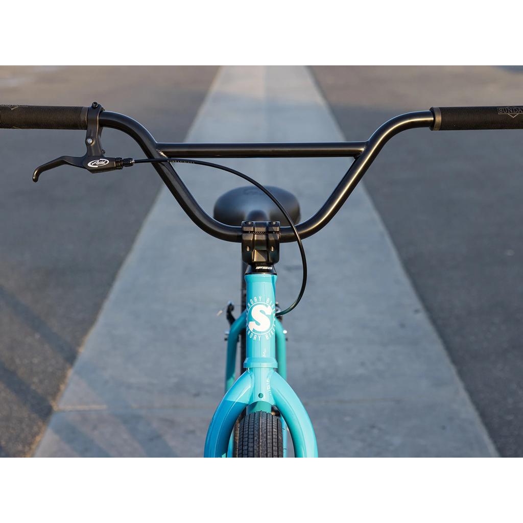 Close-up of Sunday High C 29 Inch Bike (2025) in blue, highlighting the handlebars and front tire on a sunny paved path. Its cruiser geometry ensures a relaxed ride for your next adventure.