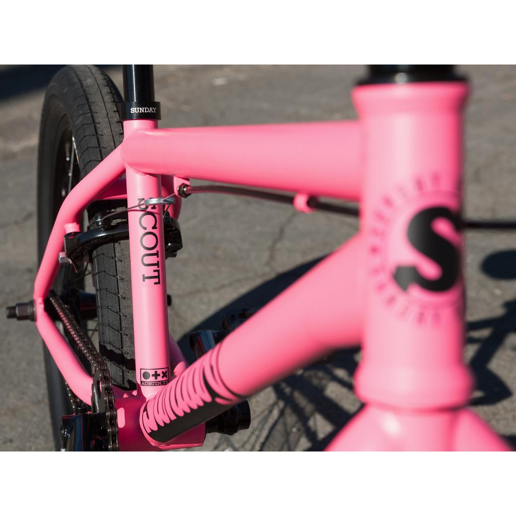 Close-up of the Sunday Scout 20 Inch Bike in bright pink, showcasing its sturdy chromoly frame, Odyssey parts, "SCOUT" label on the front fork, and Sunday logo on the frame, parked on pavement.