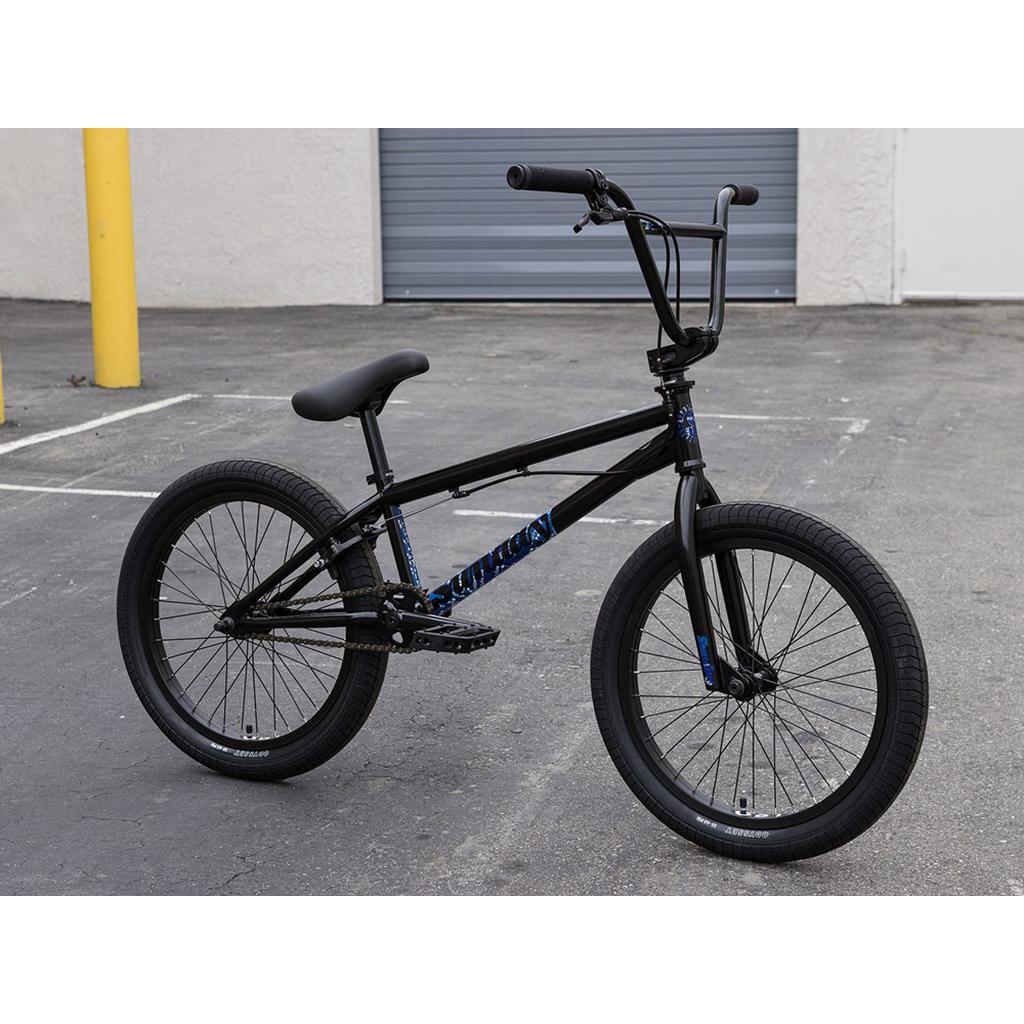 A Sunday Forecaster Maca Grasset 20 Inch Bike, black with thick tires and blue decals, is parked on an empty asphalt lot near a gray garage door.