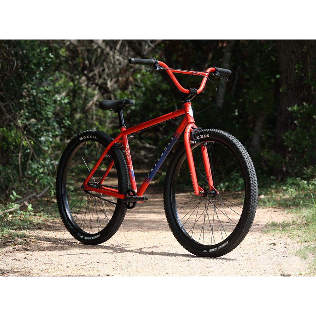 A Fairdale Taj 27.5 Inch Bike (2023) with thick tires stands on a dirt path among greenery and trees, showcasing BMX commuter style and single speed simplicity.