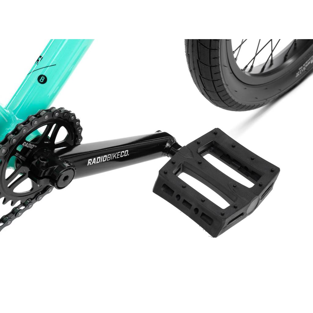 Close-up of a black RADIOBIKECO. crank, chainring, and pedal beside the rear wheel on a Radio Saiko 18 Inch Bike kids BMX with Salt Tracer Tyres.