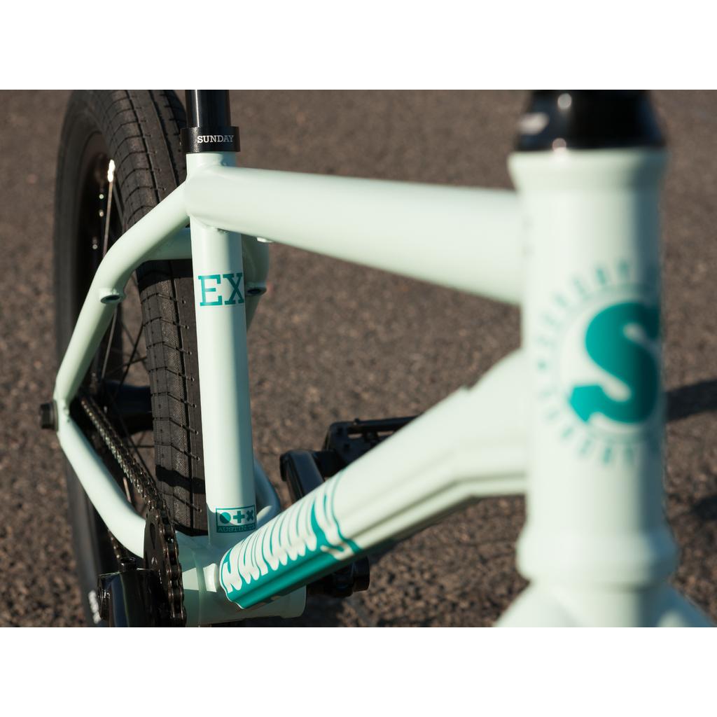 Close-up of a light green Sunday EX Julian Arteaga 20 Inch Bike with black tires, featuring the "EX" label and "S" logo on the front. It has a 100% chromoly frame and is shown resting on an asphalt surface.