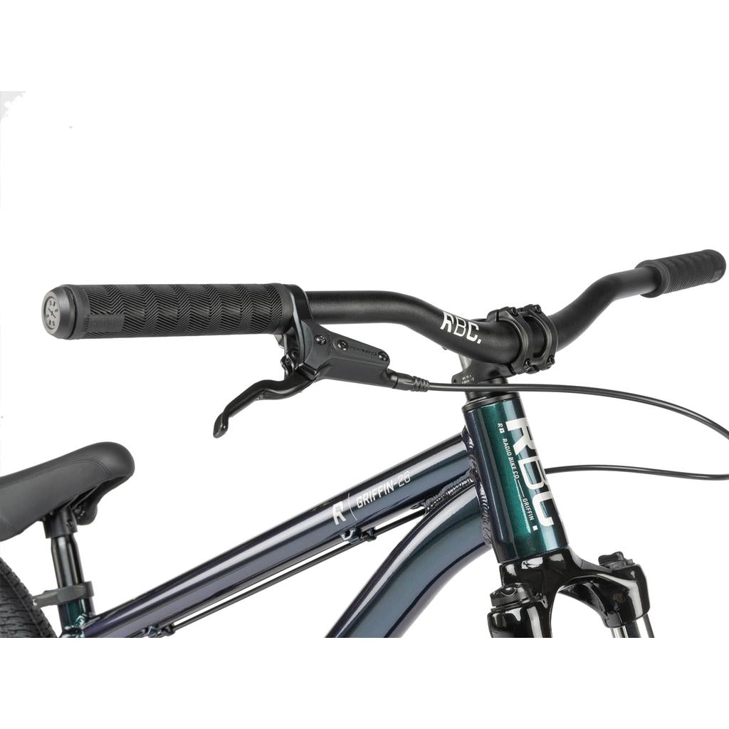 Close-up of the Radio Griffin 26 Inch Bike’s handlebars, brake lever, and front frame, showing handle grips, cables, Tektro disc brakes, and part of the front suspension fork.