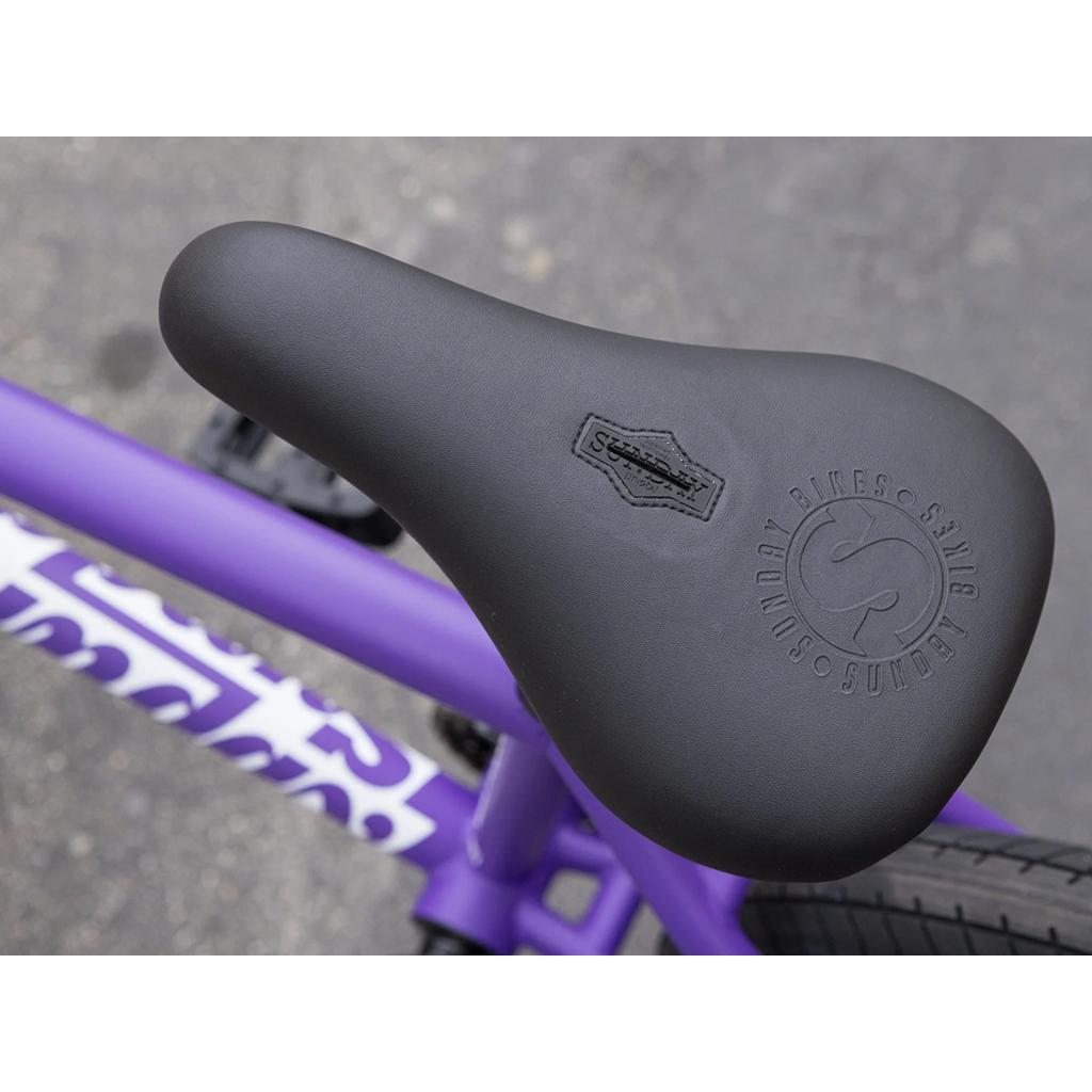 Close-up of a black seat with embossed branding on a purple chromoly frame from the Sunday Forecaster 20 Inch Bike, showing part of the tire and logo.