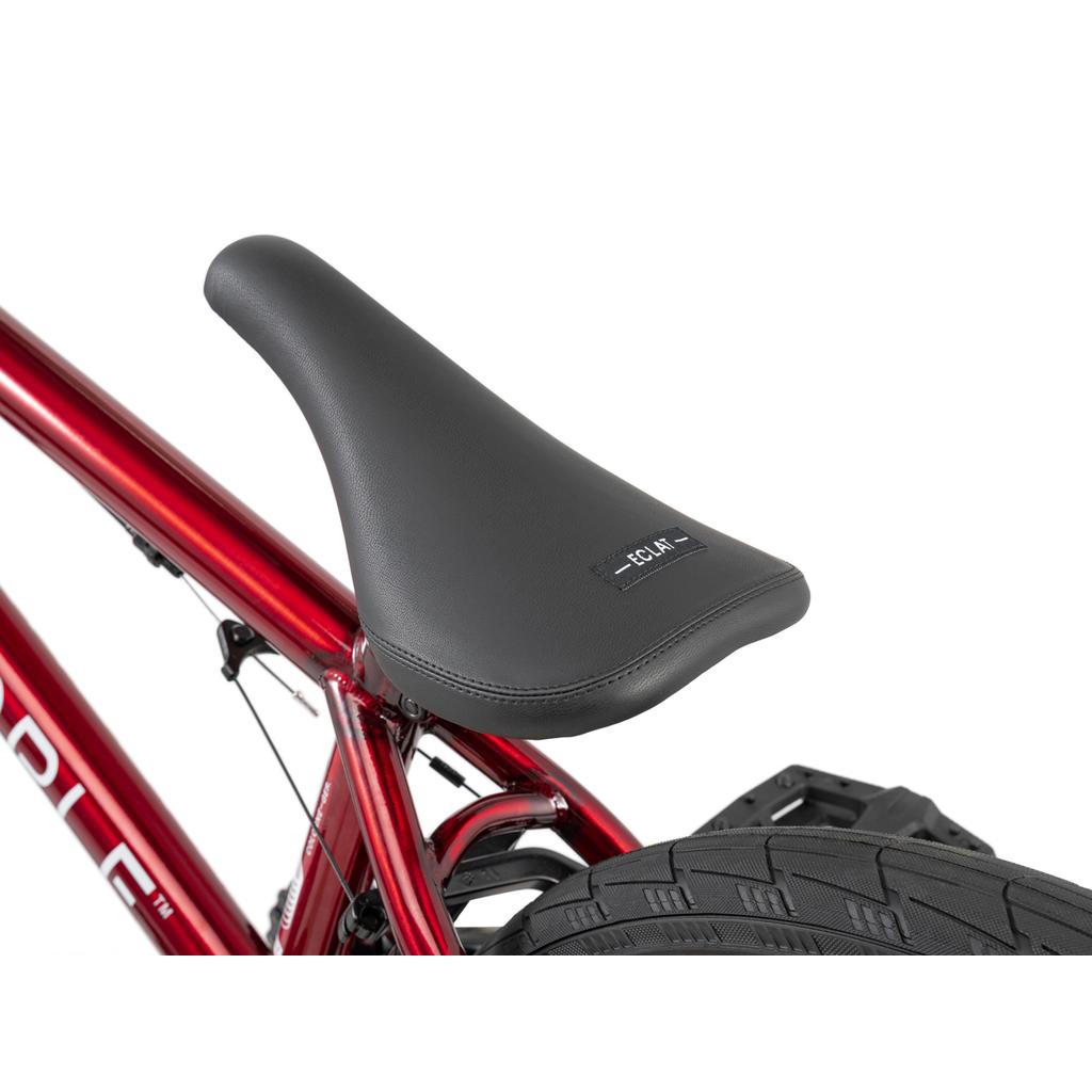 Close-up of a black ECILA saddle mounted on the Wethepeople Versus 20 Inch Bike's red frame, with part of the rear tire visible—ideal for skatepark BMX enthusiasts.