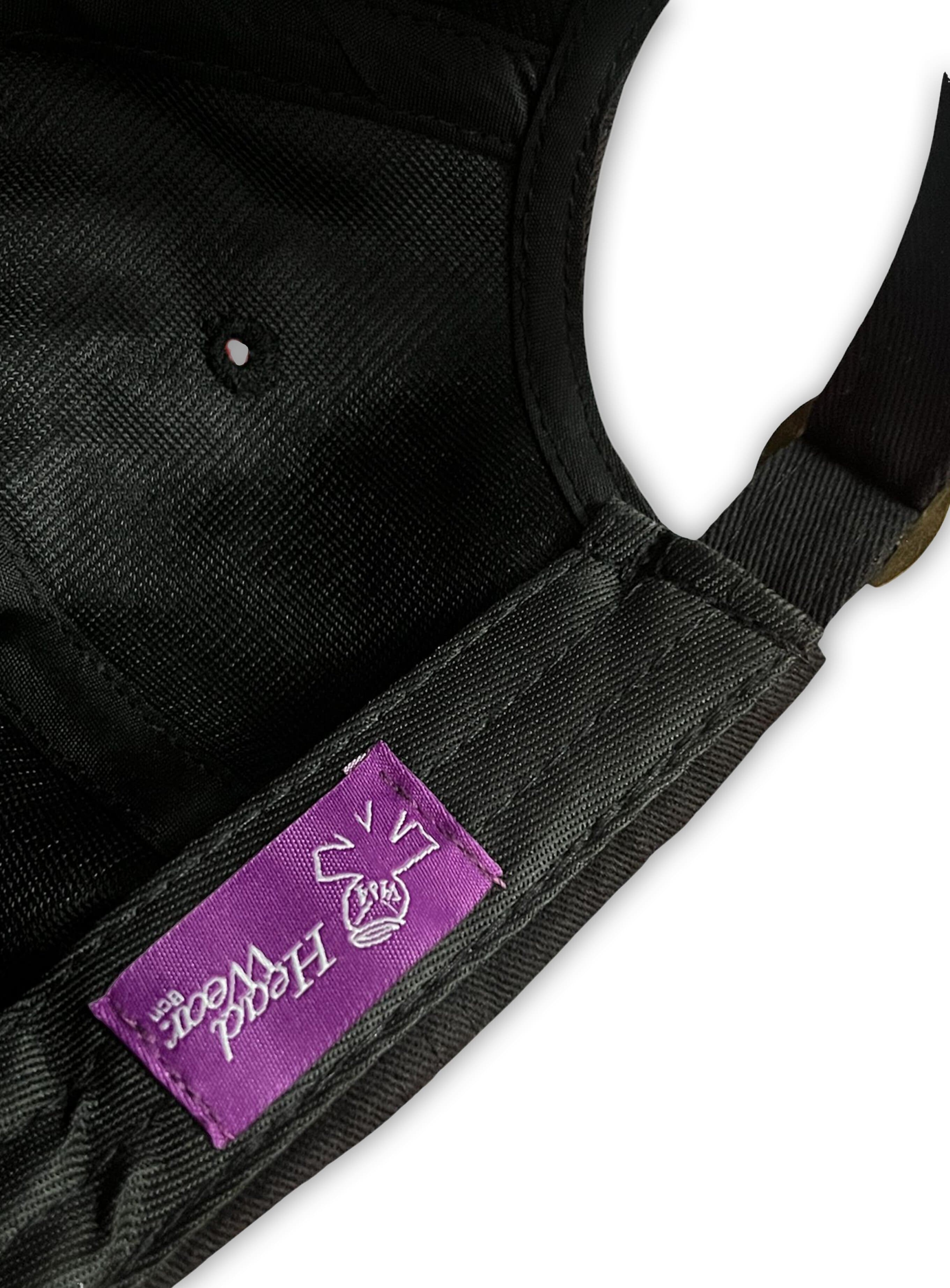 Close-up of the V1da The Dad Cap, a black unstructured hat with a purple stitched label featuring a white "HOOD WOOD" graphic and the word "BASK.