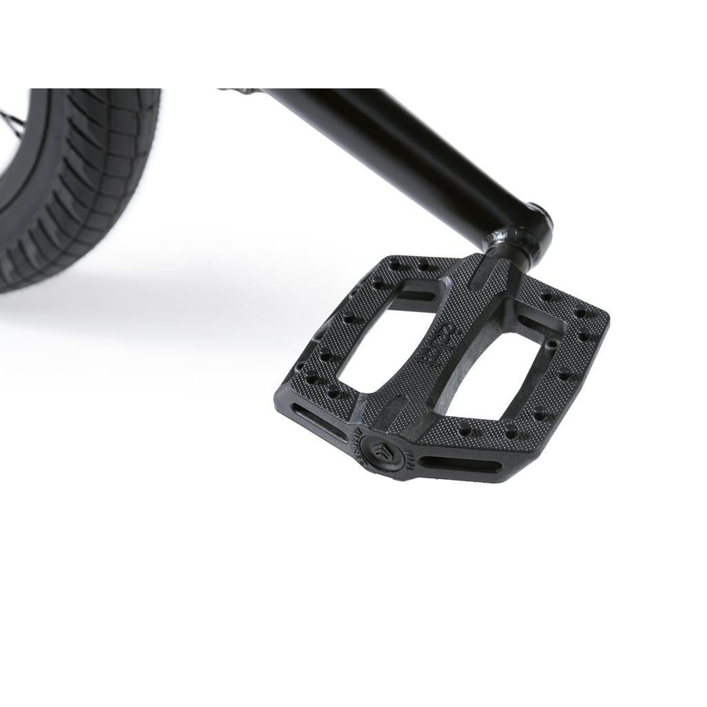 Close-up of a black bicycle pedal attached to the crank arm on a Wethepeople Revolver 20 Inch Bike, with part of the rear wheel and tire visible in the background.