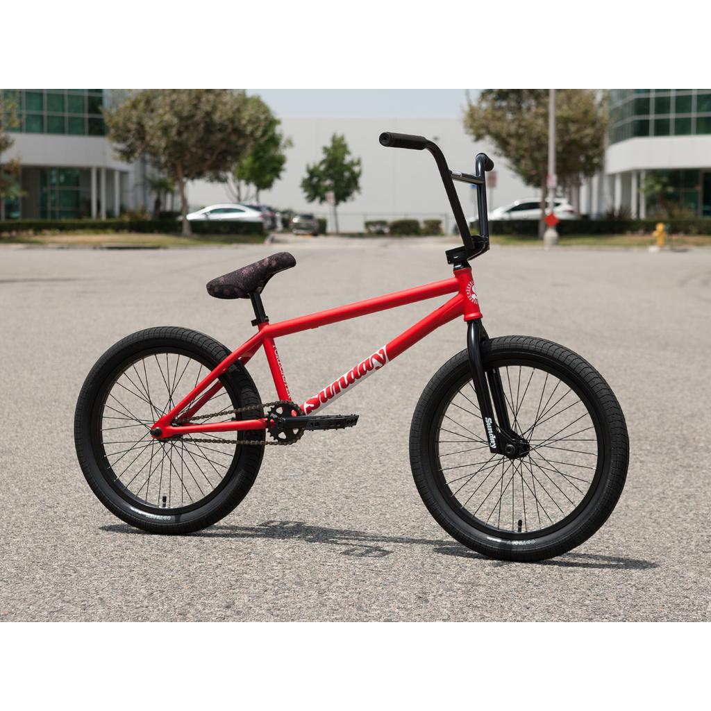 A Sunday Forecaster 20 Inch Bike (2025) / Brett Silva in red, featuring black tires and handlebars, is parked on a paved road in an empty parking lot with buildings and trees visible in the background.