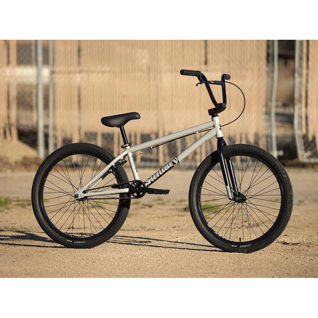 A Sunday Model C 24 Inch Bike with a matte gray chromoly frame, black tires, seat, and handlebars is parked on dirt in front of a chain-link fence.