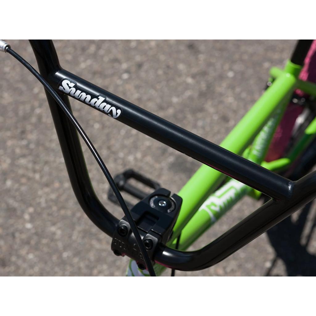 Close-up of the Sunday Blueprint 20 Inch Bike (2023) shows a black BMX handlebar, green frame with "Sunday" branding, and Odyssey Springfield brakes—ideal for both beginners and advanced riders. Photographed outdoors.