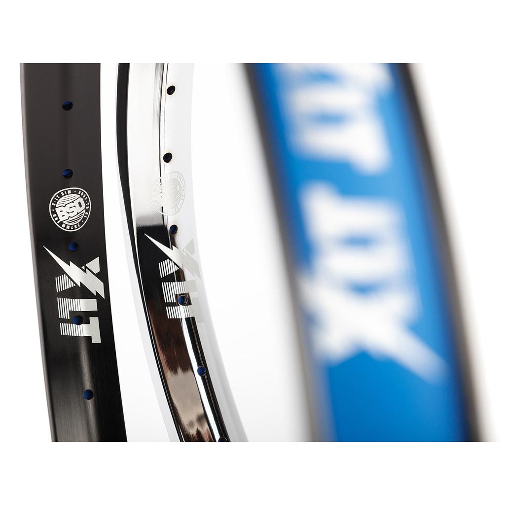 Close-up of two BSD XLT Rims—one black, one silver—with visible branding, plus part of a blue rim blurred in the foreground.
