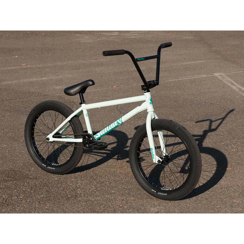 A light blue Sunday EX Julian Arteaga 20 Inch Bike with a 100% chromoly frame, black handlebars, seat, and tires is parked on empty asphalt.