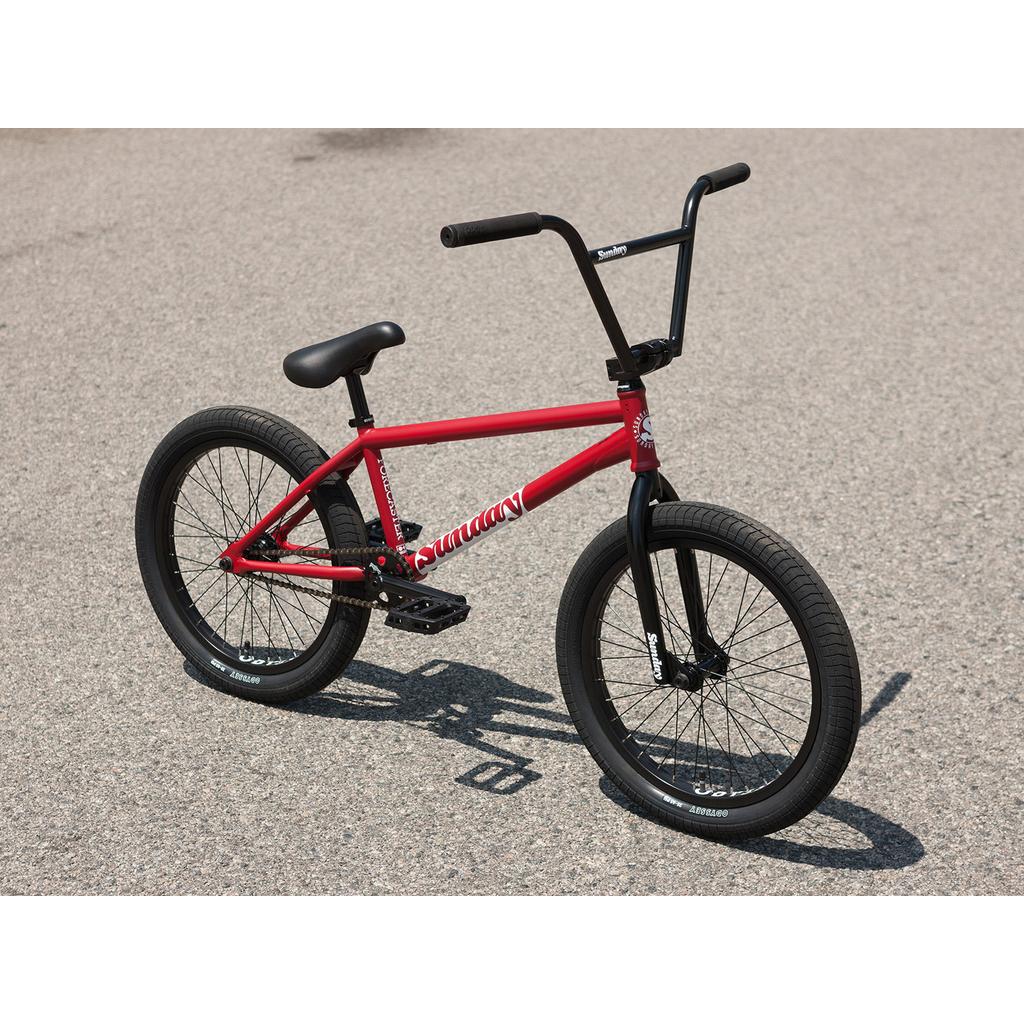 The Sunday Forecaster 20 Inch Bike, featuring a red chromoly frame with black tires, handlebars, and seat, is parked on an asphalt surface.