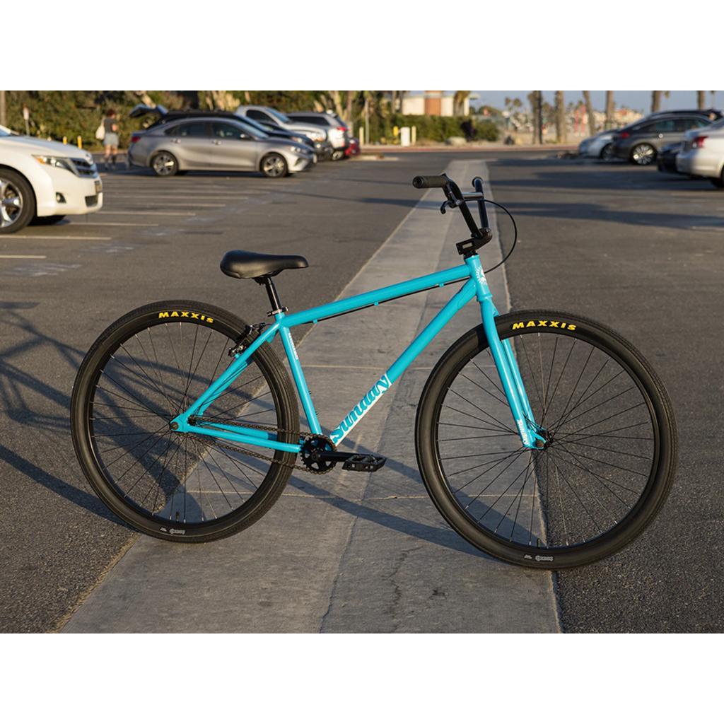 A Sunday High C 29 Inch Bike (2025) with blue frame, cruiser geometry, and black tires is parked on a concrete path in a lot, with cars and palm trees visible in the background.