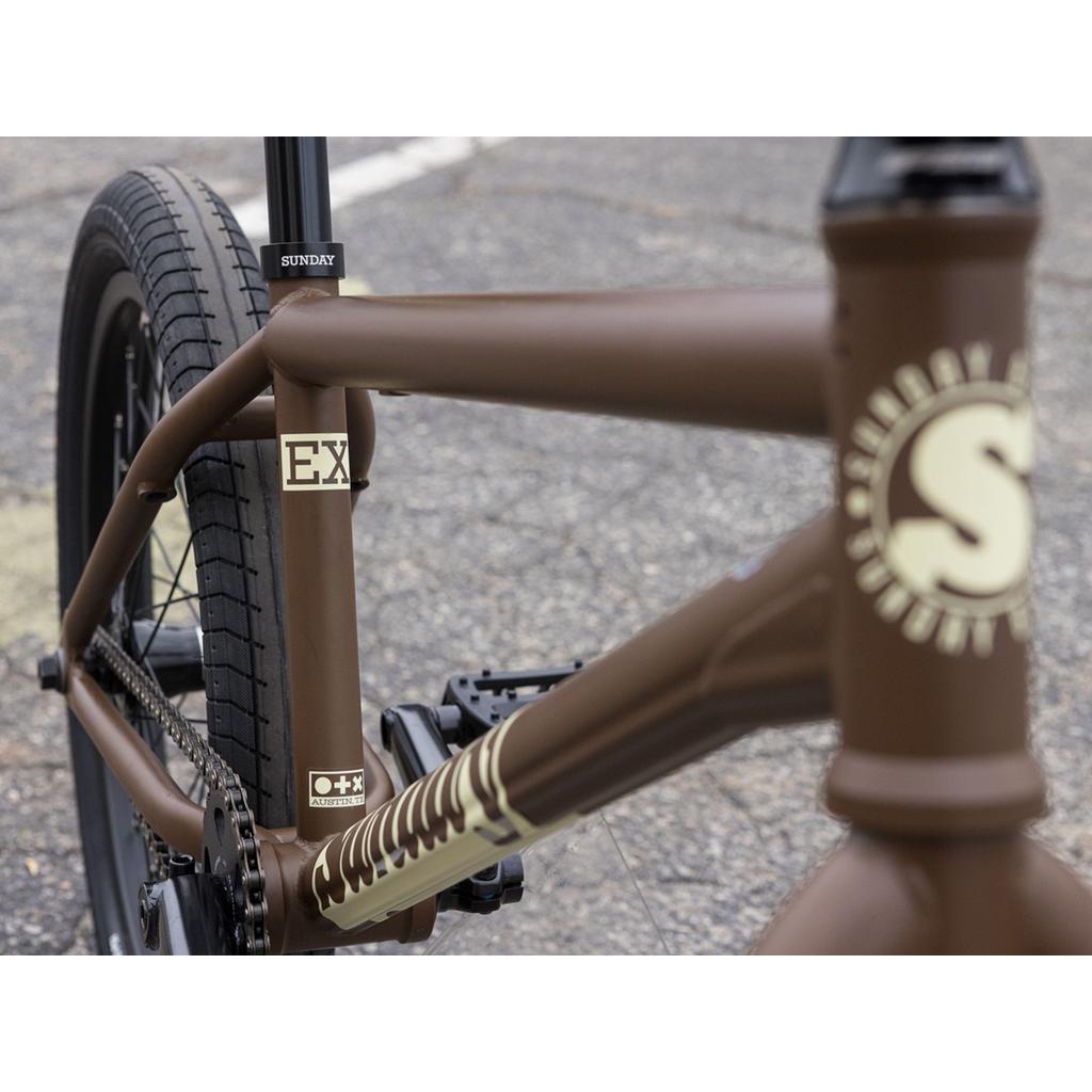 Close-up of a Sunday EX Julian Arteaga 20 Inch Bike featuring "Sunday" and "EX" branding, brown chromoly frame, rear tire, chain, and pedal visible on asphalt—a Julian Arteaga signature BMX.