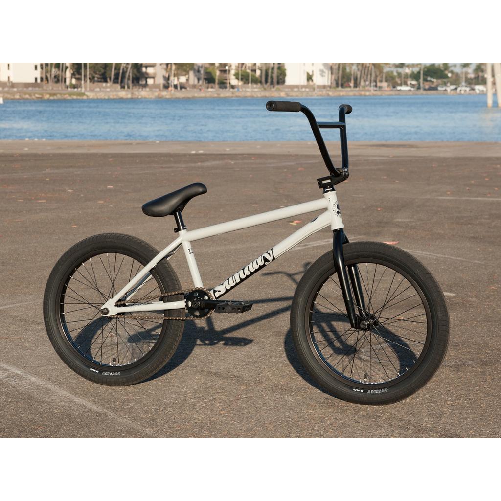 A Sunday EX Erik Elstran 20 Inch Bike featuring a 100% chromoly frame and thick tires is parked on an empty paved surface near a body of water.