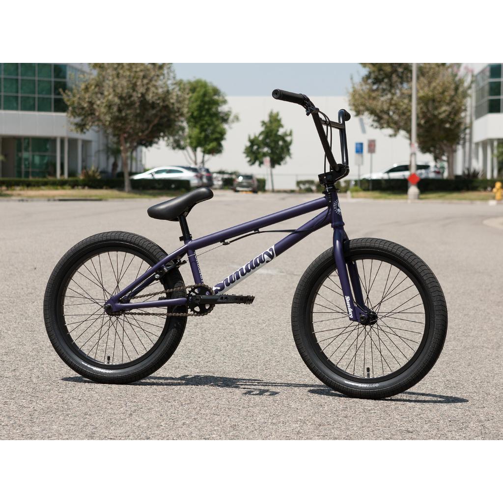 A Sunday Forecaster Maca Grasset 20 Inch Bike in purple with black tires and a gyro setup is parked on a paved street beside modern office buildings and trees.
