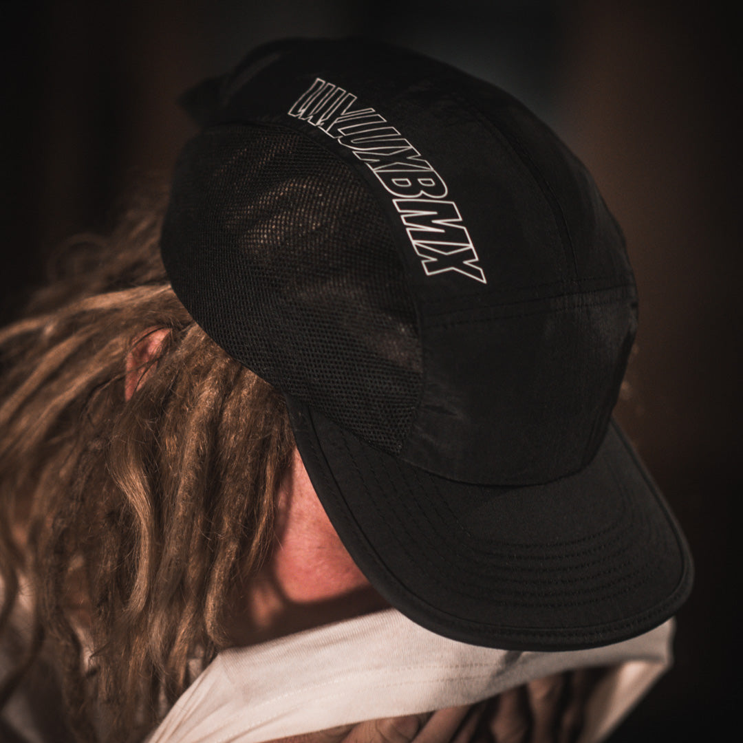 A man with dreadlocks holding a hat while browsing through a selection of LUXBMX Bike Athletics Caps - Black at a BMX store.