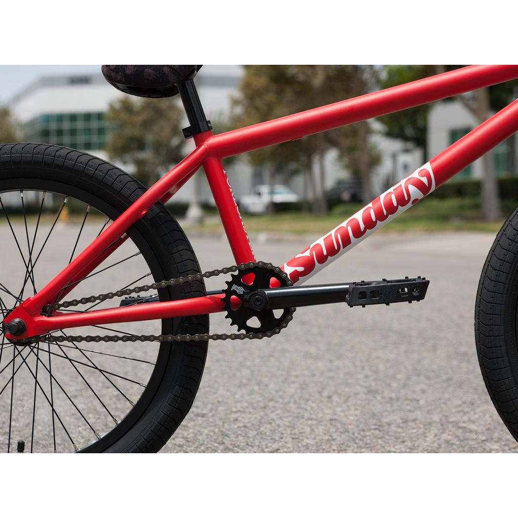 Close-up view of the drivetrain and rear wheel of a red Sunday Forecaster 20 Inch Bike (2025) / Brett Silva with a 100% chromoly frame, photographed outdoors on pavement with blurred buildings and trees behind.