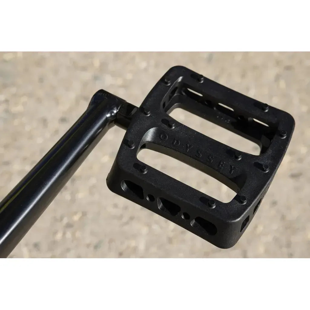 Close-up of a black BMX pedal with "Odyssey" embossed on it, attached to a black crank arm, highlights the quality parts featured on the Sunday Soundwave Special Bike (2025) against a blurred outdoor background.