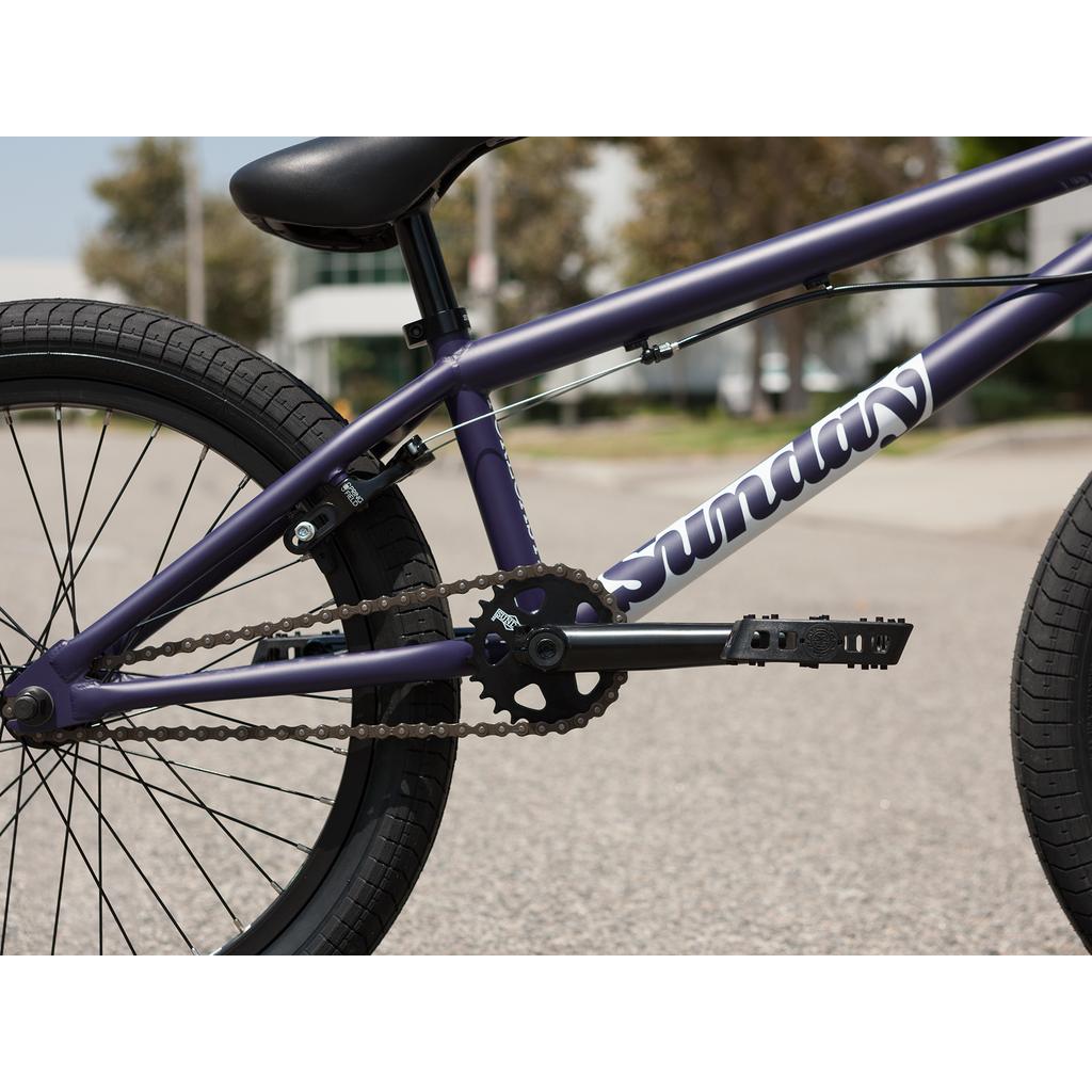 Close-up of the drivetrain and rear wheel of the purple Sunday Forecaster Maca Grasset 20 Inch Bike, showing "Sunday" branding on the frame and a gyro setup, photographed outdoors on pavement.