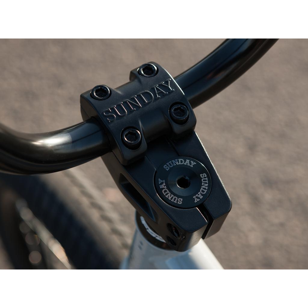 Close-up of a black handlebar stem with "SUNDAY" engraved, mounted on a white 100% chromoly Sunday EX Erik Elstran 20 Inch Bike frame, photographed outdoors.